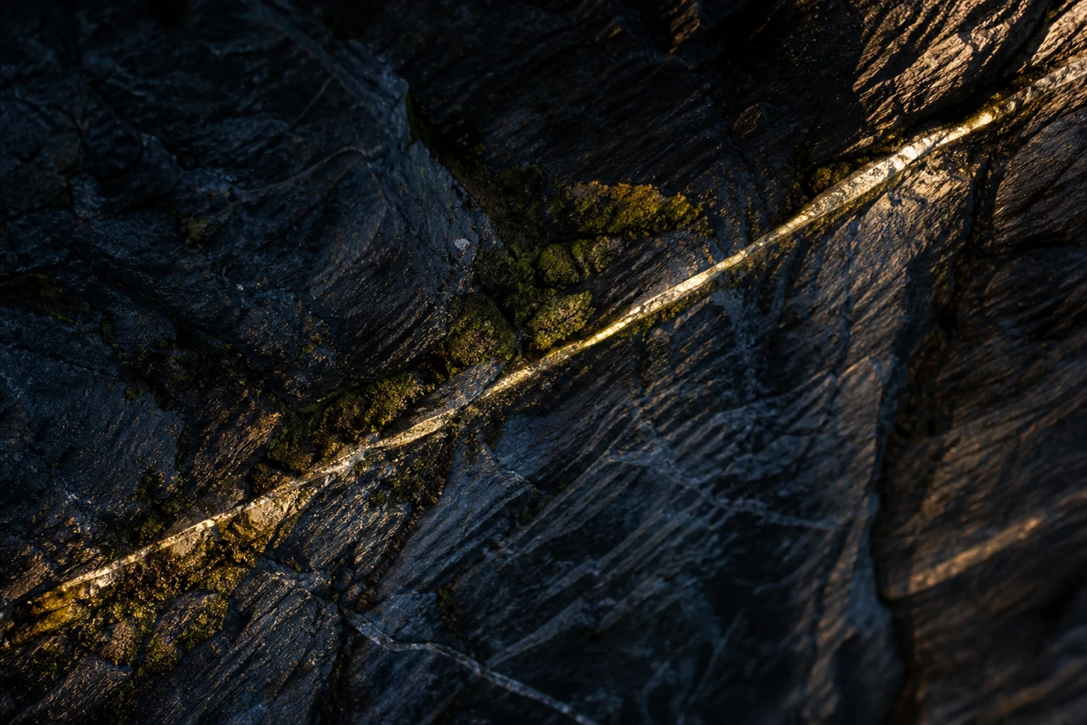Surface de pierre sombre avec une fine fissure lumineuse et des mousses discrètes.